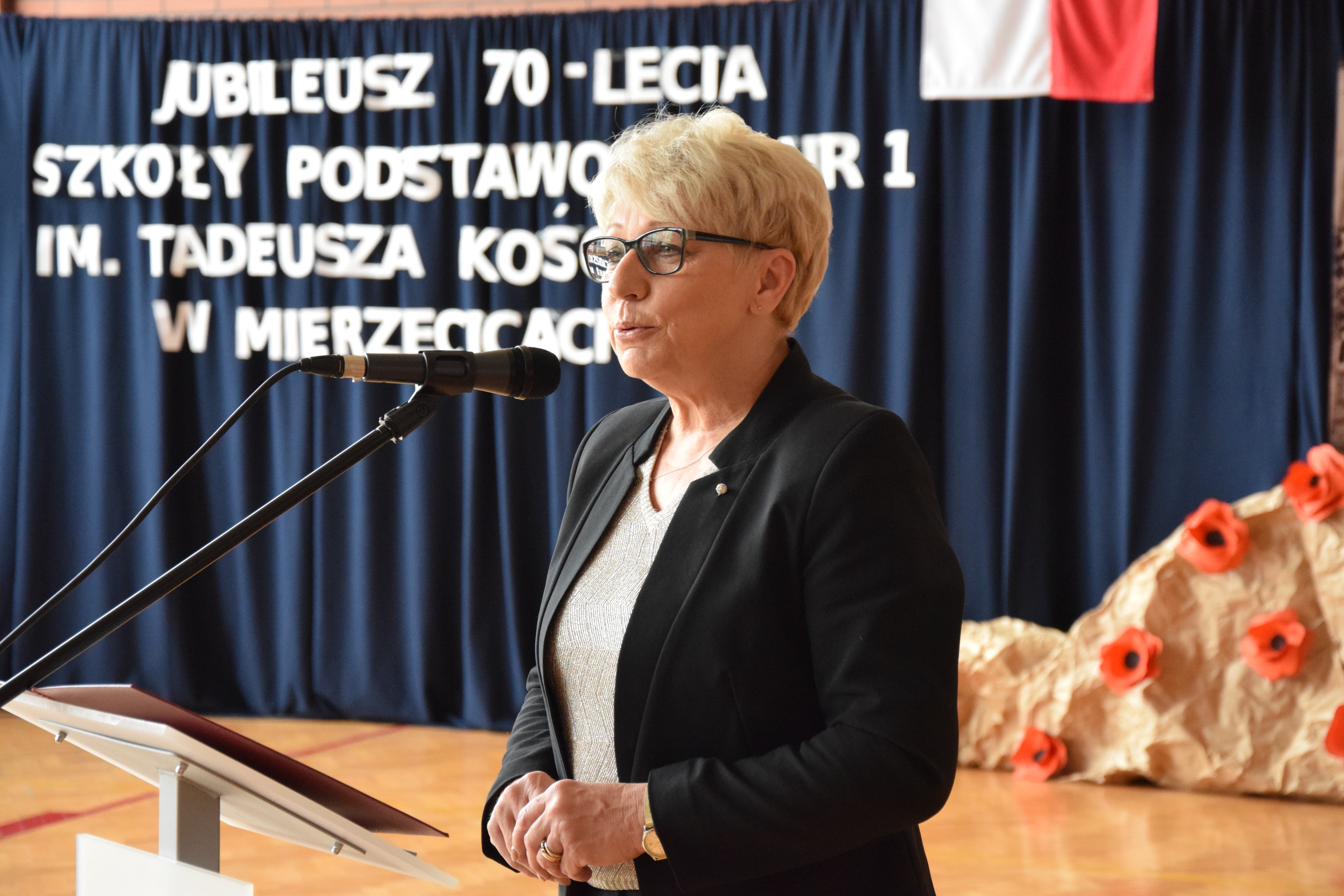 Zdjęcie przedstawiające przemawiającą byłą panią dyrektor Bożenę Jach na tle dekoracyjnego napisu "Jubileusz 70-lecia SP1"